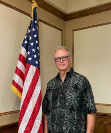 John Nichols wearing a black and grey button up shirt standing next to an American flag